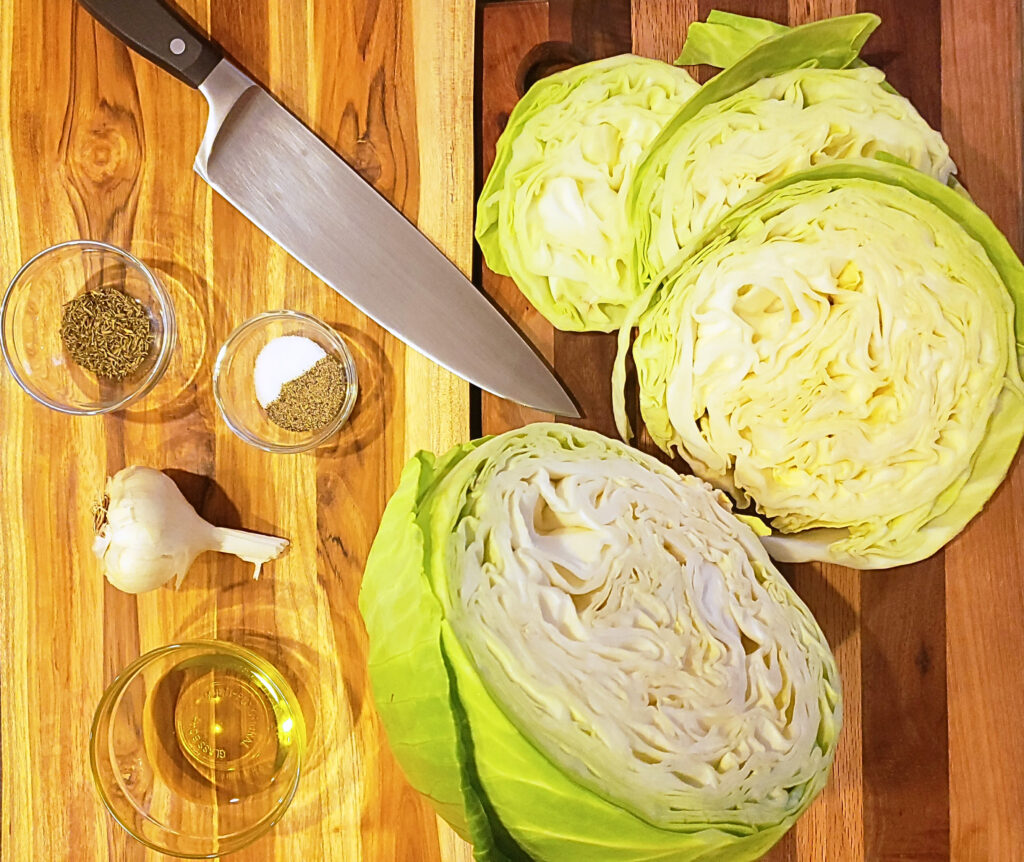 Ingredients for cabbage steaks including cabbage, salt, pepper, thyme, garlic, and olive oil on a wooden cutting board with a knife