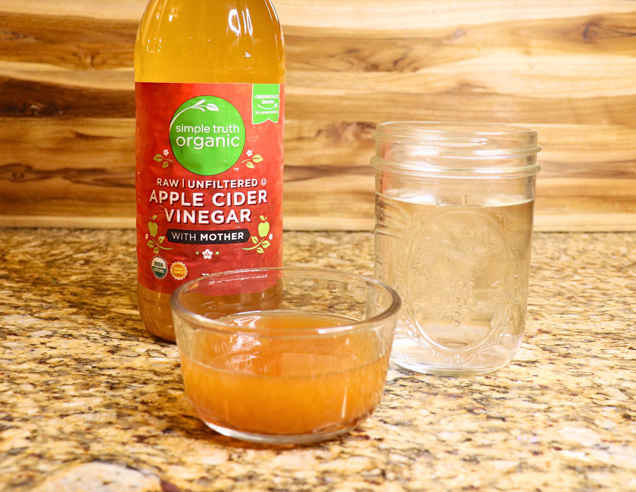 A bottle of Apple cider vinegar next to two glass jars with white vinegar and ACV sitting on a counter
