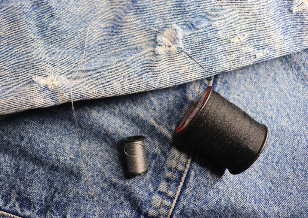 Denim clothing with a hole that has a threaded needle going through it and two spools of thread