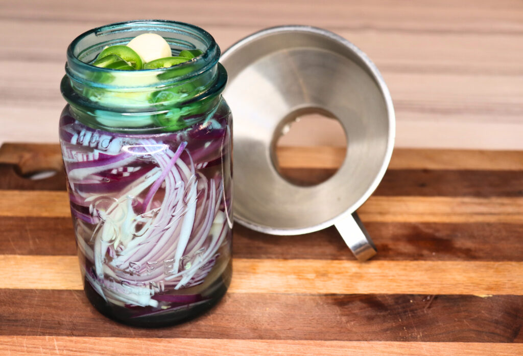 Mason jar filled with pickled spicy onion ingredients by a canning funnel