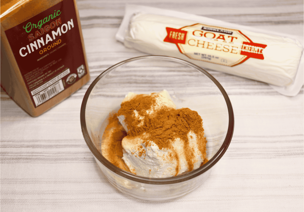 A glass bowl with goat cheese, honey, and cinnamon next to a log of cheese and a cinnamon bottle.