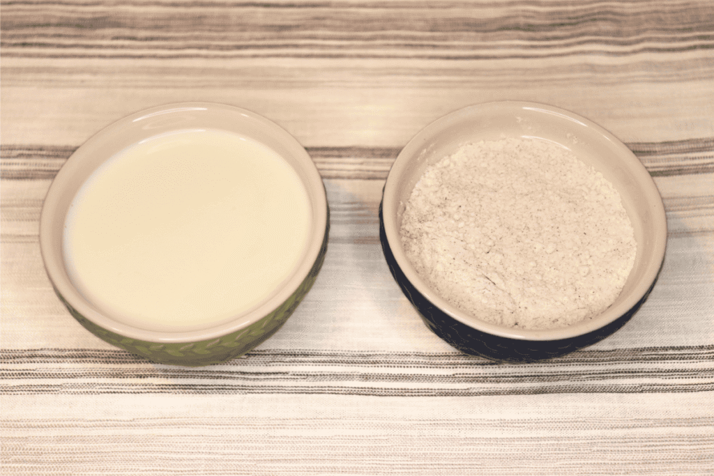 A bowl of milk next to a bowl of flour and seasoning on a white and gray table cloth