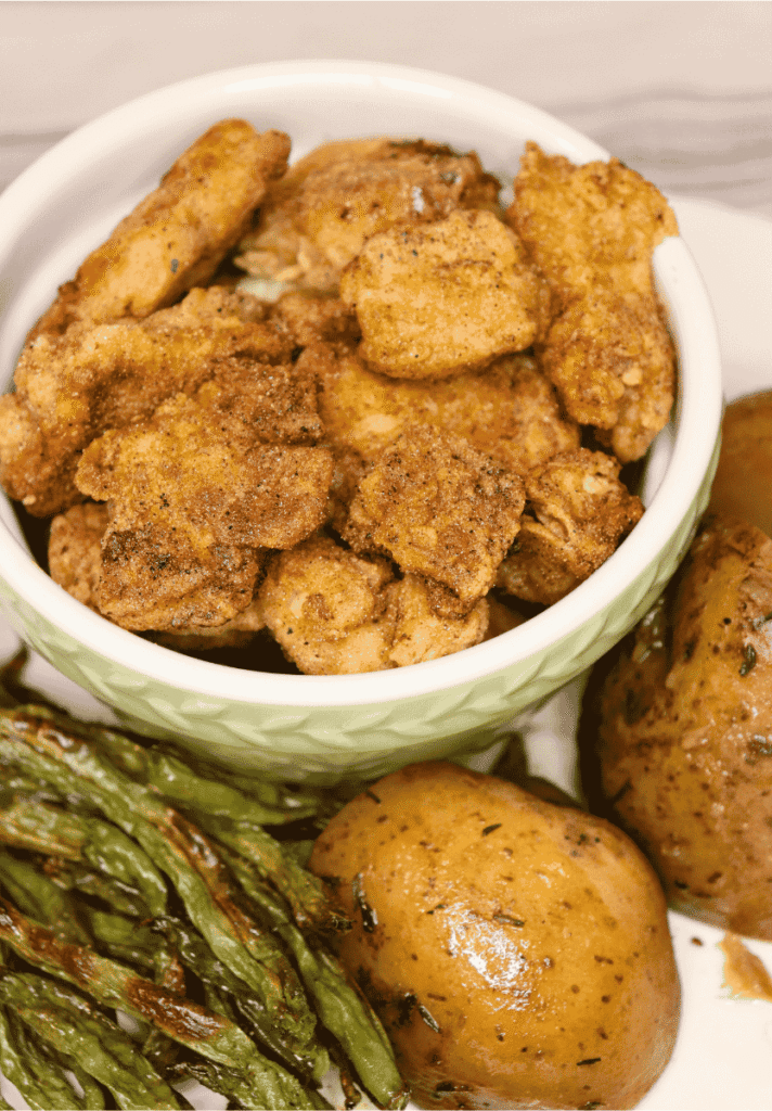 Lightly breaded homemade chicken nuggets in a bowl on a plate with roasted potatoes and green beans