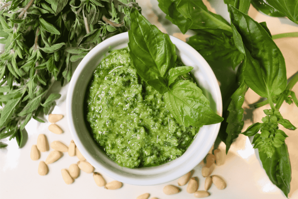 Basil pesto in a white bowl next to fresh basil leaves, fresh oregano, and pine nuts