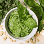 Basil pesto in a white bowl next to fresh basil leaves, fresh oregano, and pine nuts