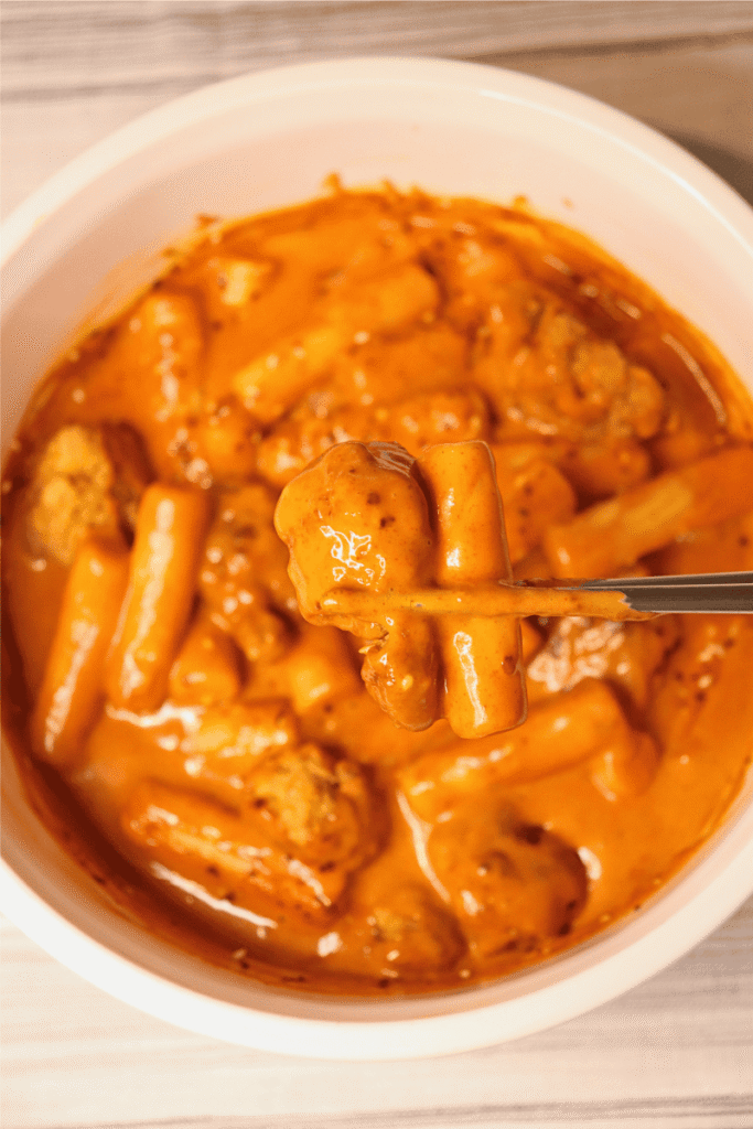 Close up of a bite of creamy tteokbokki and fried popcorn chicken being held by chopsticks in front of the full dish