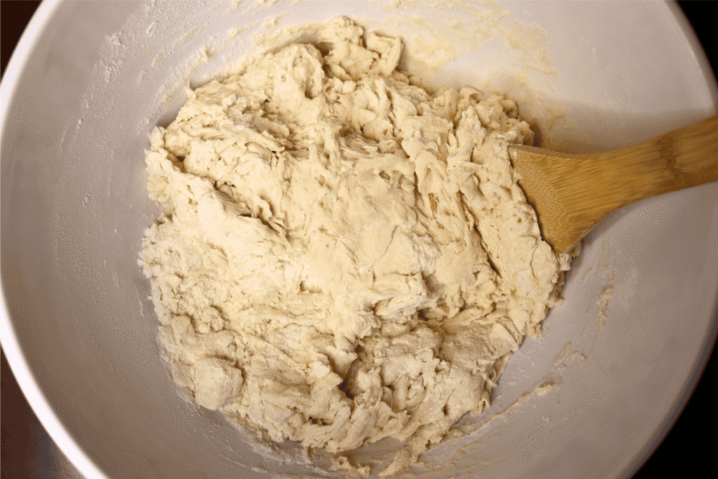 A wooden spoon mixing bread dough in a white mixing bowl