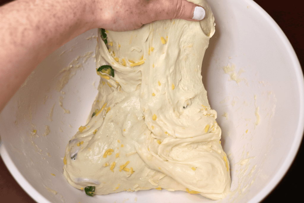 A hand stretching jalapeño cheddar sourdough bread dough upward and out of a white mixing bowl