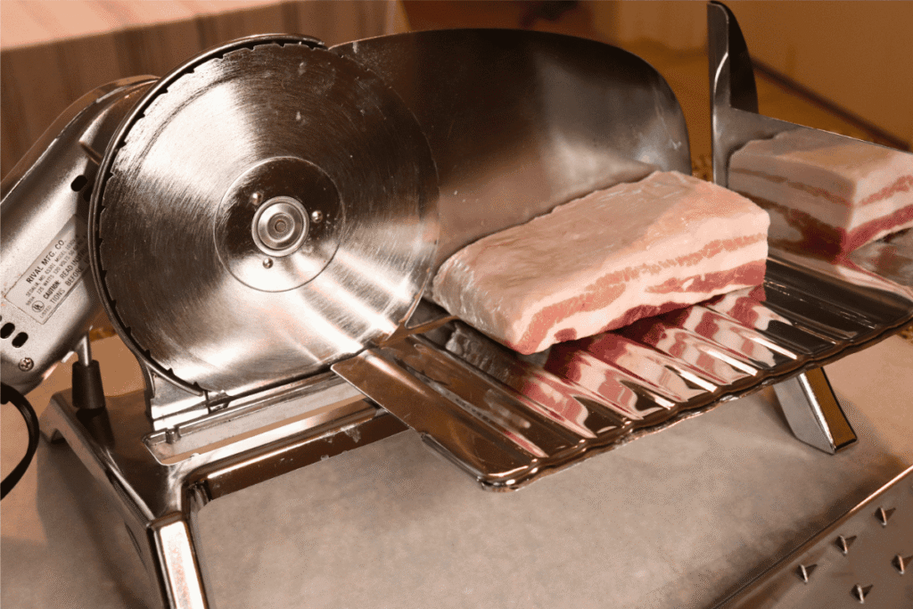 An electric deli meat slicer with raw pork belly ready to be sliced on it