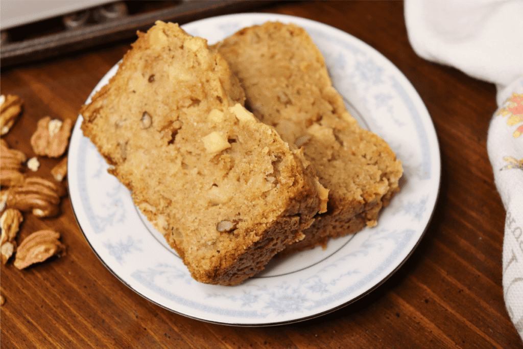 Two slices of apple bread on a saucer next to a kitchen cloth and pecans