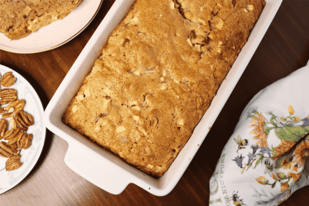 Apple bread loaf in a ceramic loaf pan by a kitchen towel and a plate of pecans